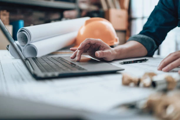 Estimator working a plan set on a laptop, hard hat and rolled drawings on the desk