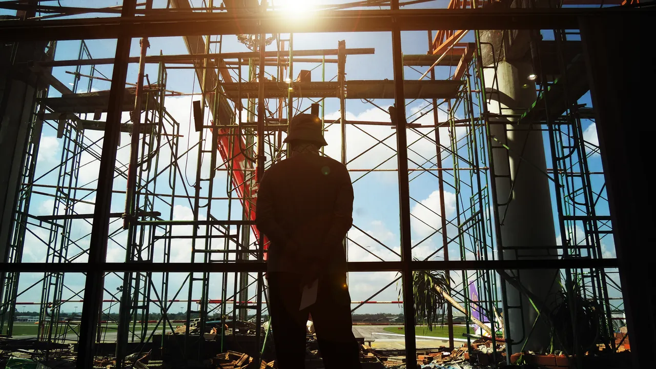 Contractor silhouetted against scaffolding on an active construction site, drawings in hand